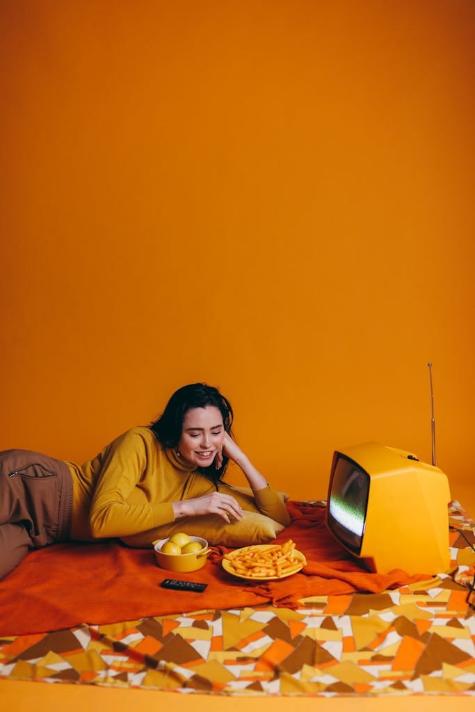 Woman enjoying a snack while watching a retro TV against a vibrant yellow background.
