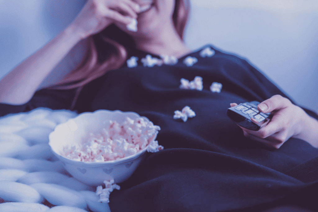 Mujer sosteniendo control remoto y palomitas mientras observa películas en servicios de streaming.